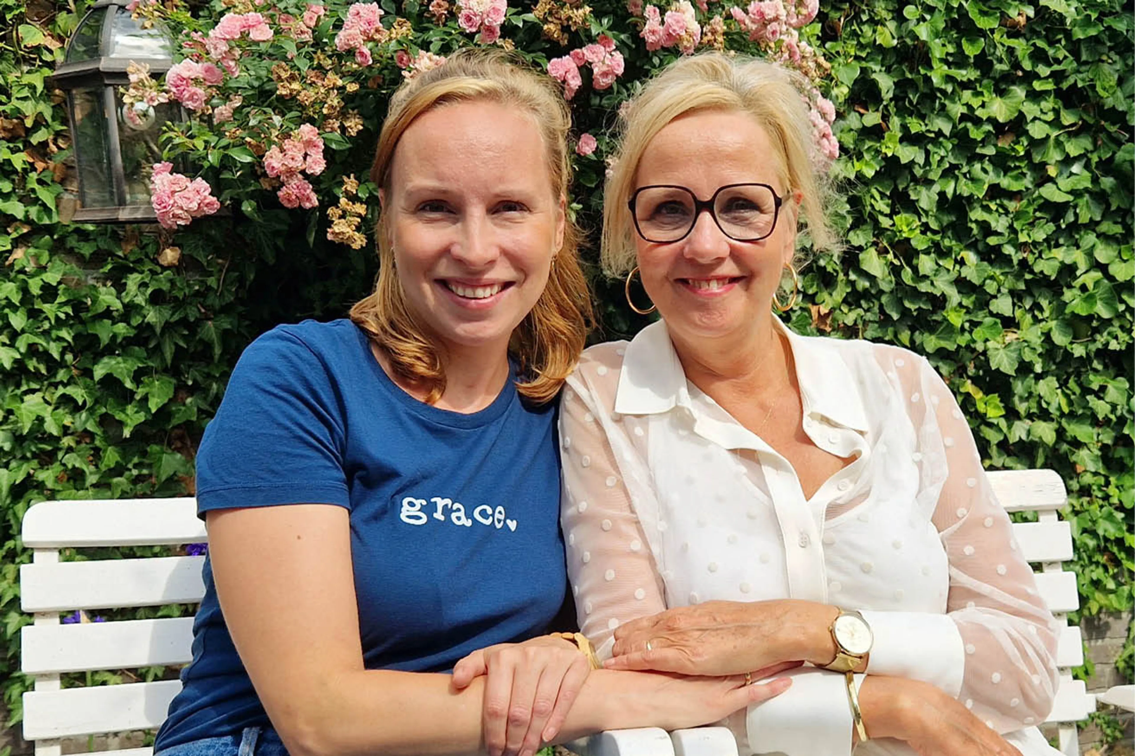 Op de foto staan Vasthi samen met haar moeder. Ze zitten op een wit bankje en achter hen is een groene heg met roze bloemen. Moeder en dochter hebben allebei blond haar en lachen. De moeder draagt een bril en heeft een witte blouse aan, Vasthi heeft een blauw t-shirt aan.